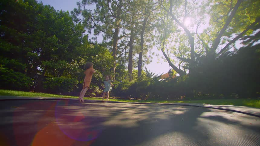 A little boy and a little girl are jumping on a black trampoline in a backyard with several trees behind
