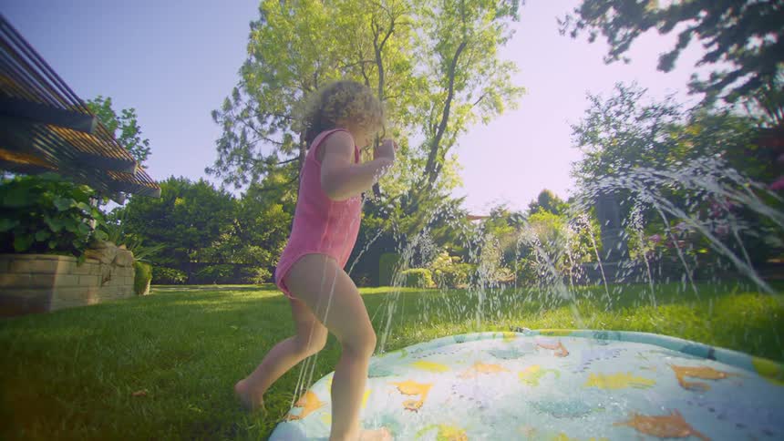 A young girl steps into a sprinkler pool in her backyard on a summer day