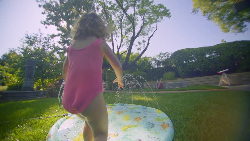 Little girl running towards a swimming pool with water jets in the middle of a backyard garden