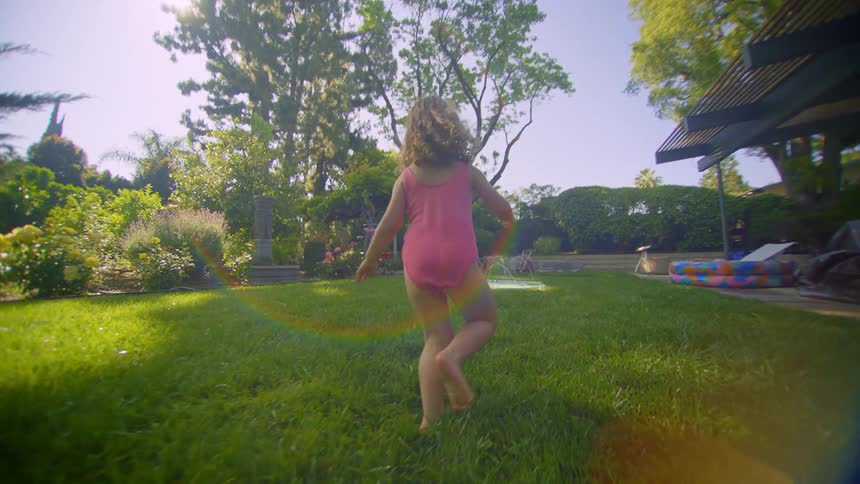 A little girl in a swimsuit is running in a grassy yard and in front of a small pool throwing water into the air