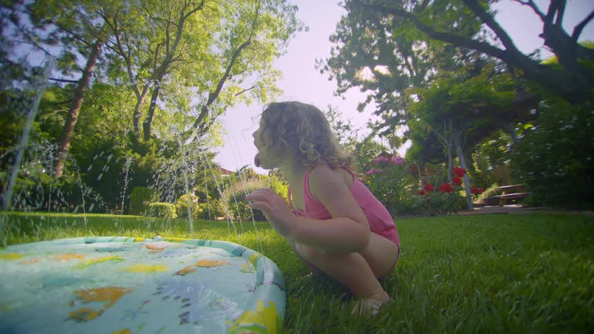 A little girl squats on the grass in a backyard with a swimming pool in front of her and she is trying to catch the water using her tongue