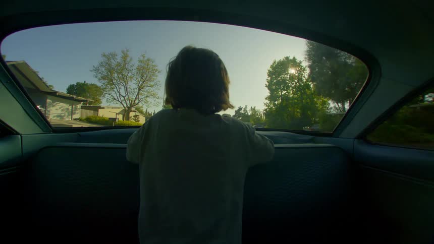 A little boy is sitting with his back on a bench looking out the window in the back of a car