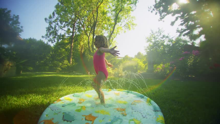 A little girl is on top of a small pool with water throwing up and she is spinning and happy and in yard with grass