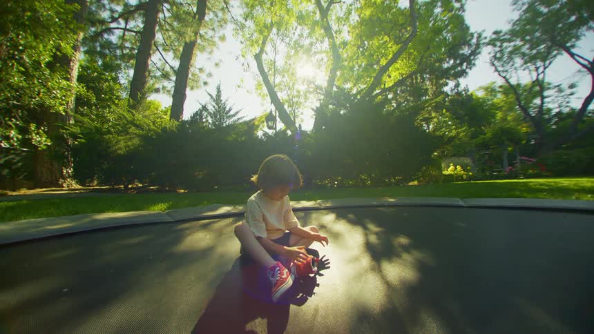 Little boy taking off his sneakers while on a trampoline in a yard surrounded by trees