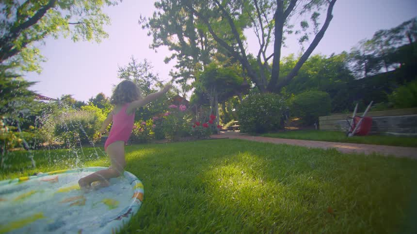 A little girl getting into a small pool in the backyard with lots of grass and she falls onto the grass and gets up