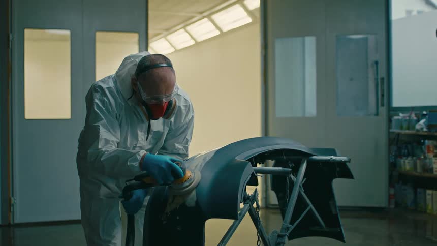 Man in protective clothing and mask polishing a part of a vehicle indoors