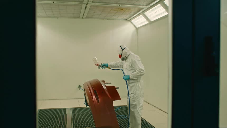 Man in protective gear using a spray gun to apply a coat to a car part inside a painting booth