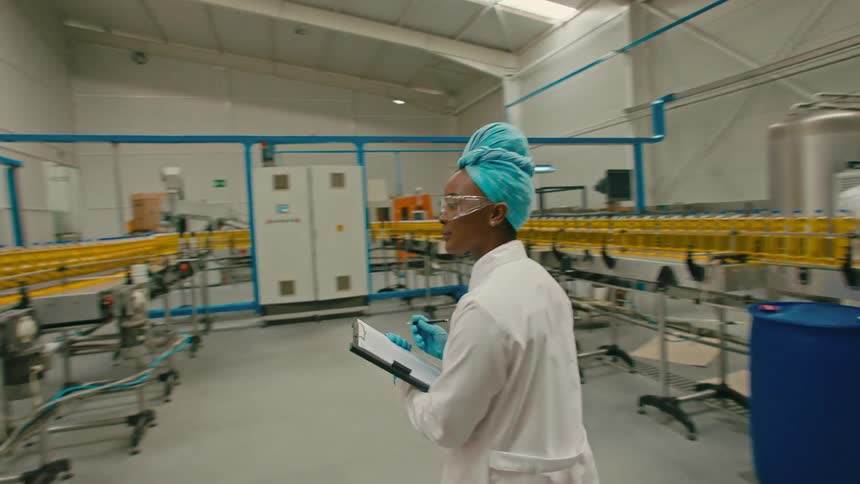 Woman taking notes on a clipboard and wearing protective glasses talking to a coworker who is analyzing the oil production process on a conveyor belt in a factory