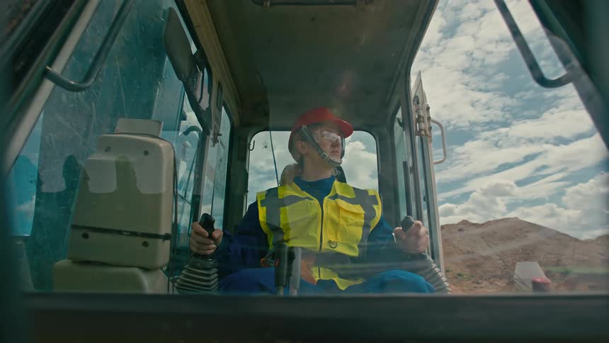 Woman in a yellow highvisibility jacket and blue helmet operating heavy machinery