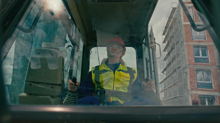 A woman in a yellow highvisibility jacket and blue coveralls sits inside a construction vehicle