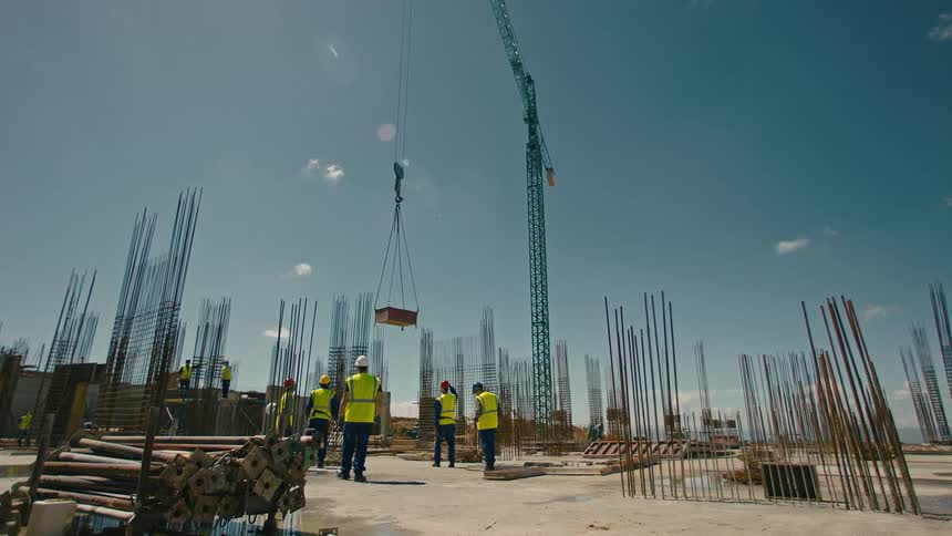 Coworkers approach building materials lowered by crane at construction site on a sunny day