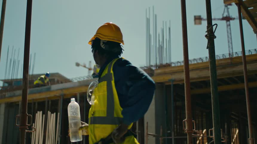A worker in a yellow vest and hard hat standing on a construction site drinking water and overseeing the progress of the building project