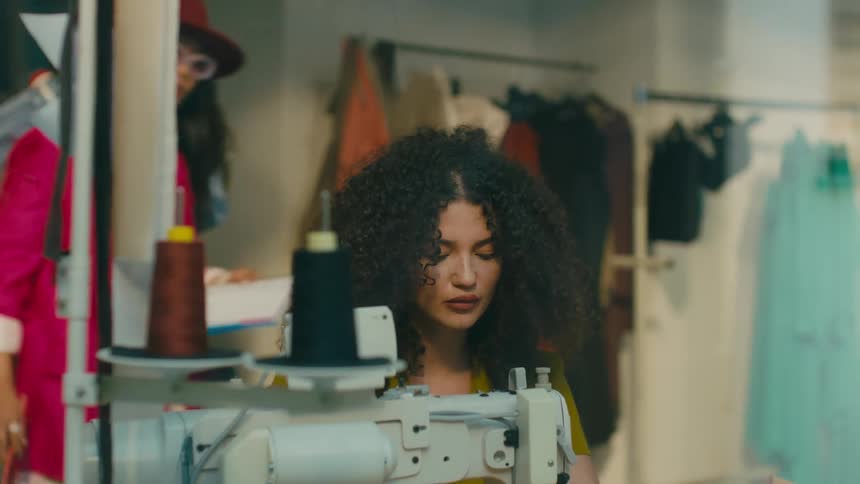 Clothing store with a woman wearing a hat and a pink blazer giving instructions to two seamstresses