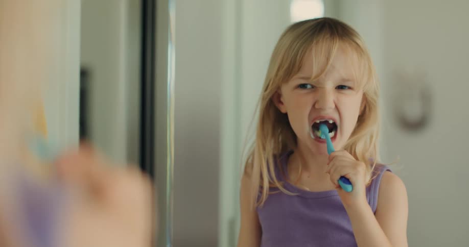 A little blonde girl brushing her teeth looking in the mirror and she smiles to see her teeth and then continues to brush them