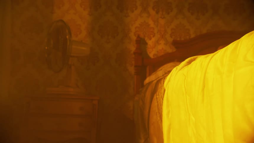 A fan spinning in a room that has strong orange lighting and there is a bed with a white cover on top