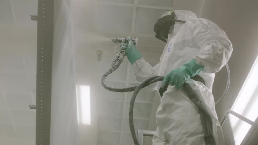 A person wearing white protective clothing is using a paint hose to paint a metal surface white