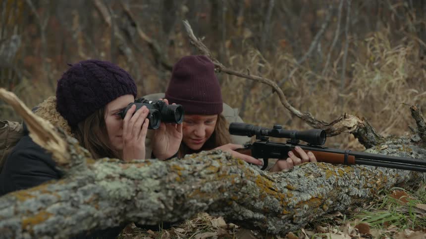 One woman aims through a rifle while another woman spots for her as they lie on the ground near the woods