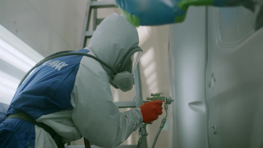 A person in white protective clothing and wearing a mask is using a paint gun to paint the door of a vehicle white