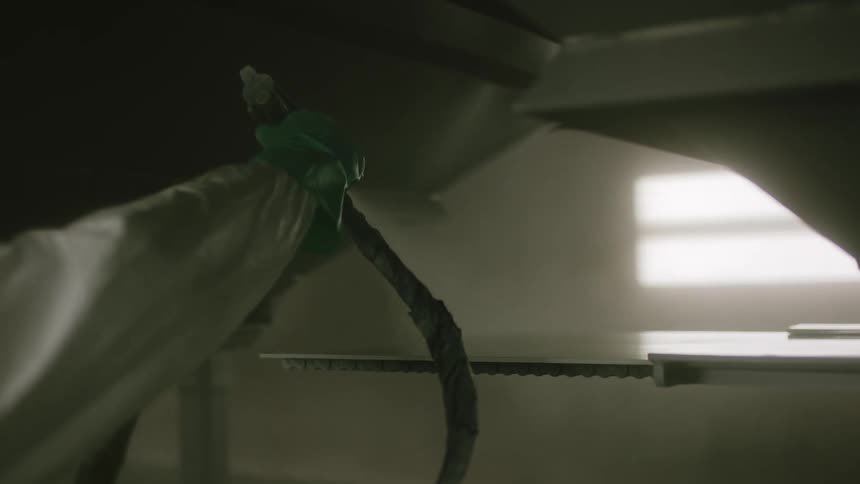 A person in white protective clothing and wearing gloves is holding a tool attached to a hose that he is using to clean a surface with jets of water