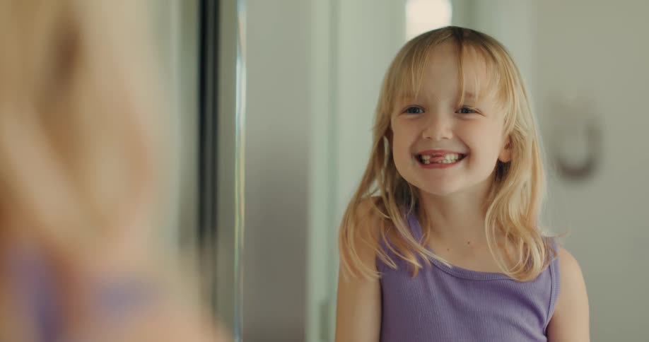 A little blonde girl is looking in the mirror and is smiling seeing that she is missing her front teeth