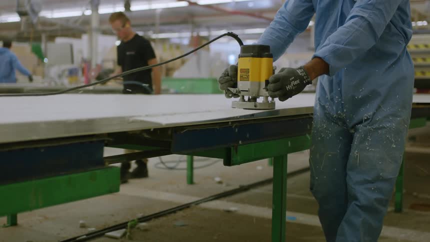 Carpenter runs a wood router along the edge of a large plank of wood while others work behind the table