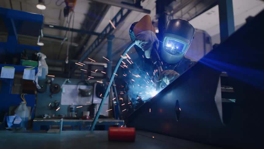 A person wearing a protective helmet is welding on a metal object and is releasing several blue sparks