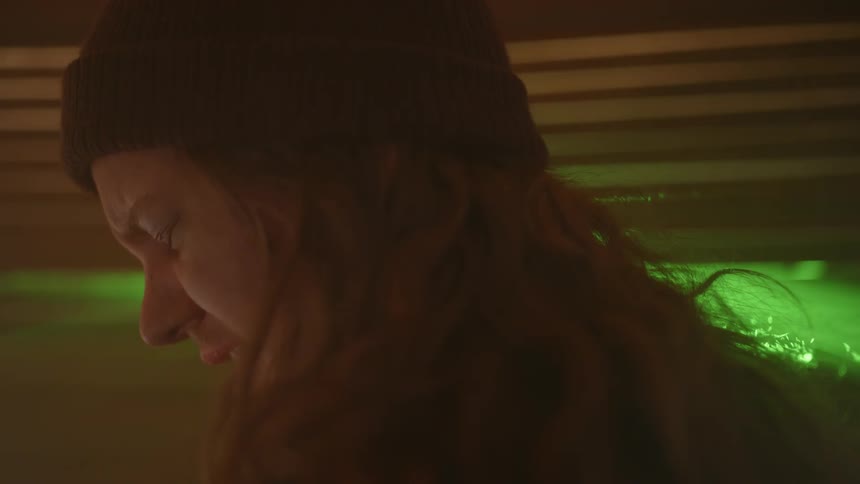 A person with long hair and wearing a cap has his eyes closed while crying and behind flashing green lights