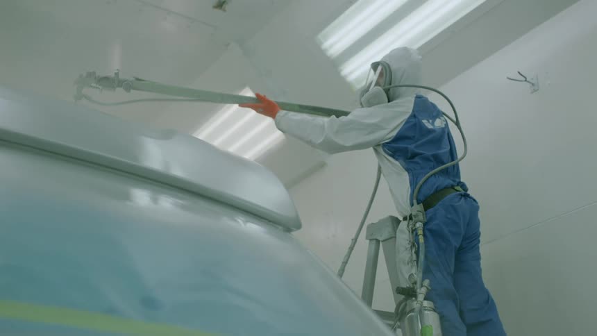 A worker stands on a ladder and sprays a metal object in a factory