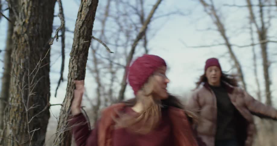 Two women wearing red hats brown coats and jeans run away in the forest
