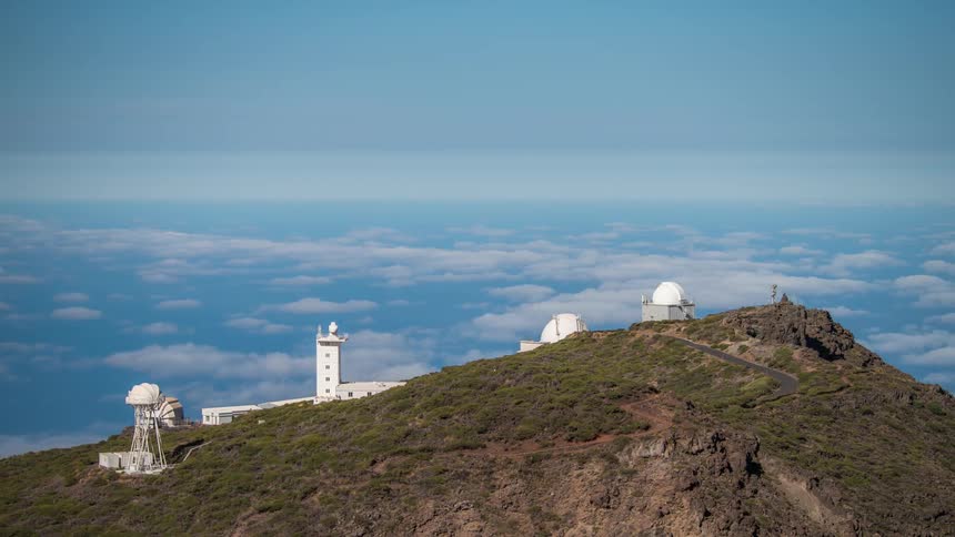 An white observatory on top of a mountain and there are several white clouds flying overhead lower than the mountain peak