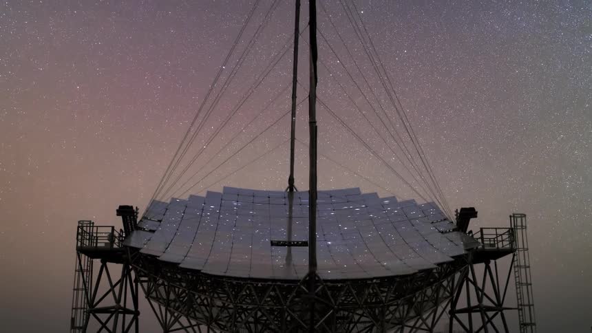 Specks of light move across a radio telescope dish at nighttime