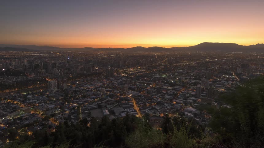 A city lights up at night as the sun sinks behind a mountain range