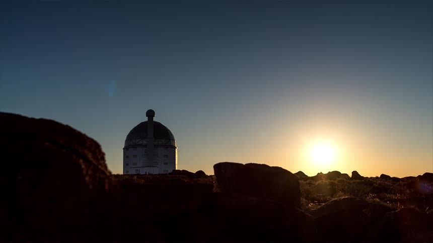 White dome of an astronomical observatory located behind a rocky terrain under a clear blue sky that turns dusk and stars appear