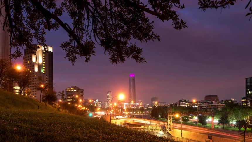 A highway with several cars passing by and the sky is pink and then it is getting dark and the city lights come on
