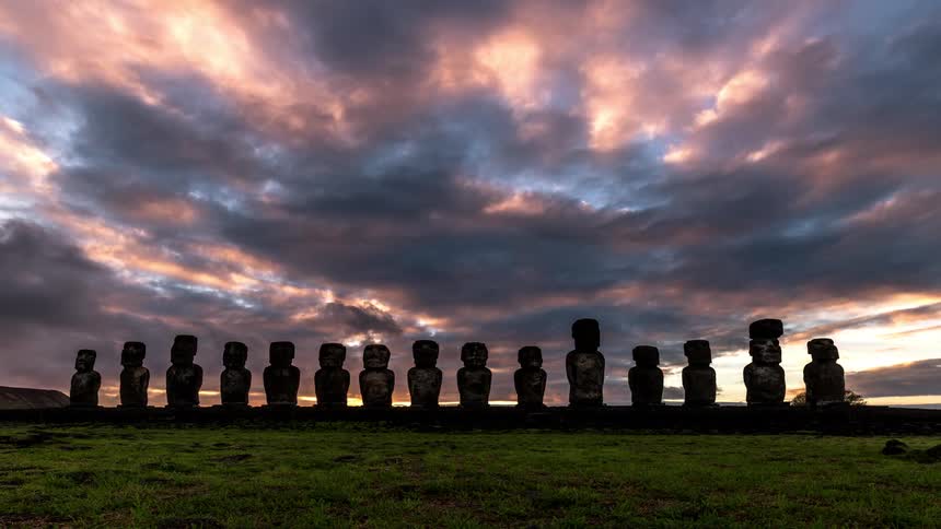 Several statues of easter island are next to each other facing forward and there are several clouds passing quickly in the sky