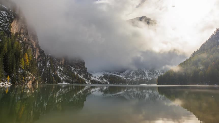 Lake reflects clouds swirling between nearby forested mountains during a winter day