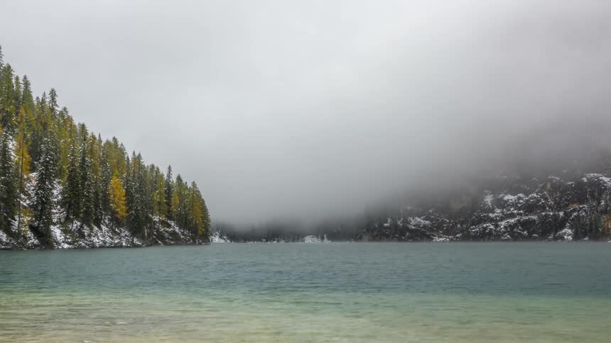 Lake with crystal clear water surrounded by trees and snowy mountains and a passing mist