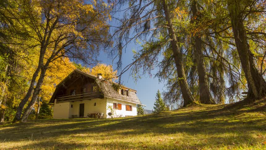 Twostory house with white walls and brown roof situated in a sloping field surrounded by trees with yellow and green leaves