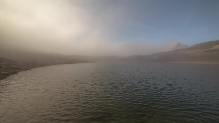 Serene lake with crystal clear waters surrounded by rocky terrain and low vegetation with majestic mountains in the background under a partly cloudy sky and a passing fog