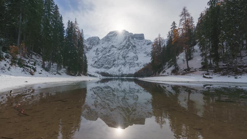 Forested mountain slopes border a lake reflecting the sun topping a snowy mountain during winter
