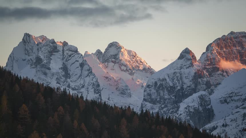 Dark clouds creep in over a snowy mountain range and a colorful pine forest