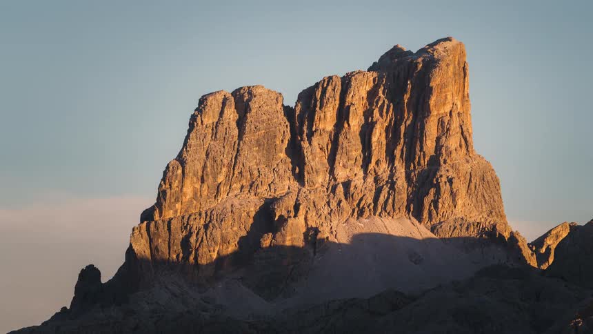 A tall rock formation is illuminated by sunlight and several clouds pass by in the background