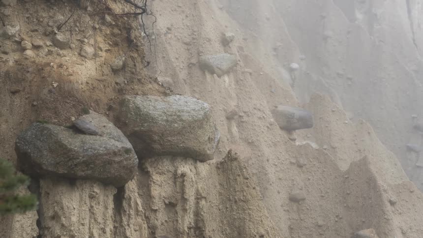 A sandy cliff with large rocks embedded within the layers surrounded in mist
