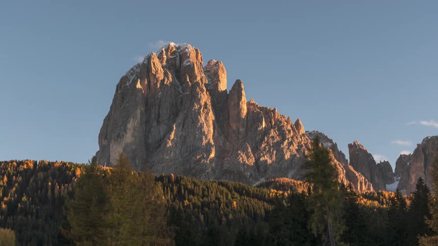 A tall rock formation in front of a pine forest and clouds pass quickly in the sky