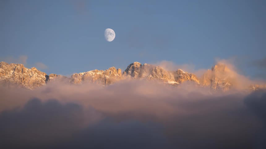 Sea of clouds passing next to mountains and the moon in a blue sky