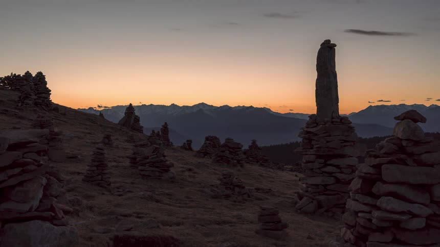 Structures made of carefully stacked rocks sit on a slope as clouds move over mountains in the distance