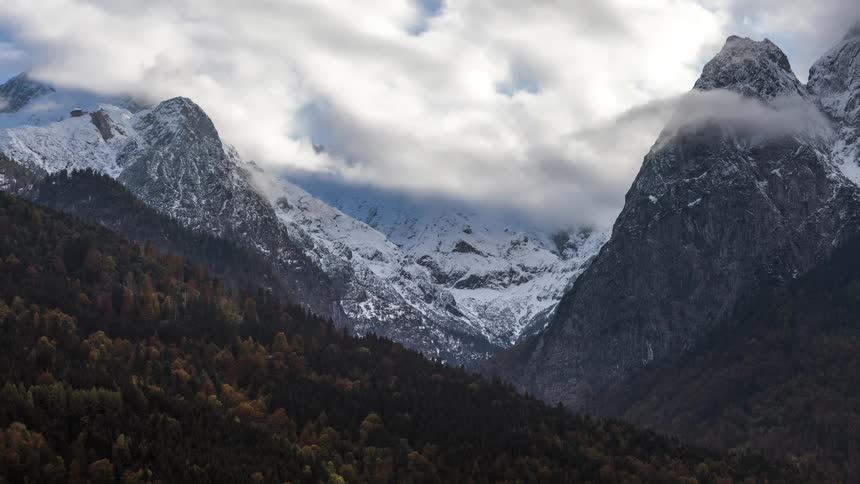 A mountain range covered in snow and in front of a mountain with a forest of trees and there are several white clouds passing in the sky