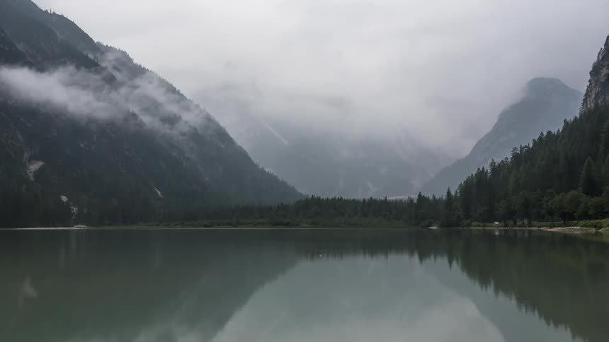Serene lake with reflections of mountains and cloudy skies framed by wooded slopes and a light mist