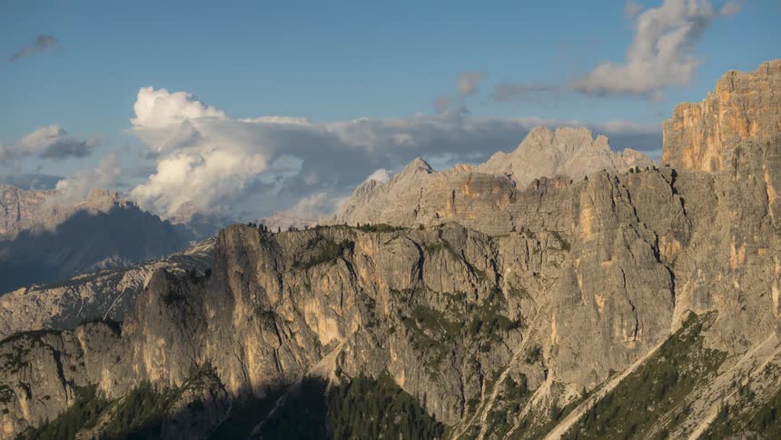 A region with stone mountains and clouds passing in the sky that are shadowing the mountains