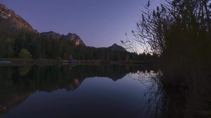 Tranquil lake at dawn reflecting the silhouette of mountains and vegetation under a gradient blue sky